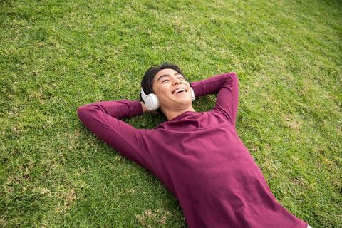 Asian man in his 20s listening to music lying on green lawn relaxing outdoors