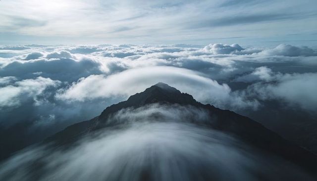 Jagged mountain peak piercing high-altitude cloud sea with lenticular dome and mist