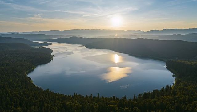 Aerial panorama of sunlit lake reflecting low sun over forested valley and distant mountains