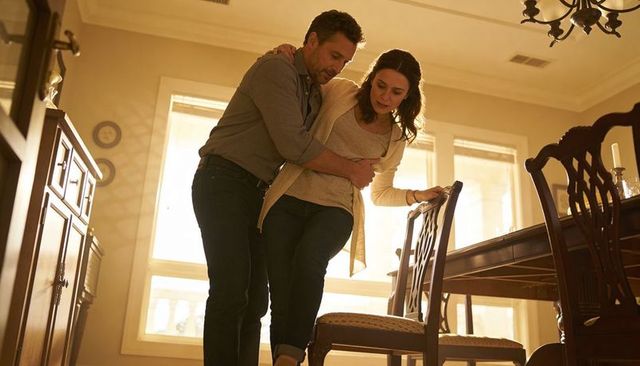 Man supporting woman rising from chair in sunlit dining room, tender caregiving couple