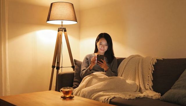 Asian woman relaxing on sofa scrolling smartphone under warm light wrapped in knit throw
