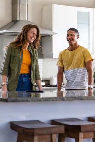 Diverse couple leaning on granite kitchen island, smiling and checking smartphone