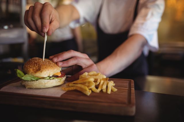 Chef placing toothpick in burger with fries on wooden board. Ideal for illustrating restaurant kitchens, culinary arts, food preparation, and professional cooking environments.