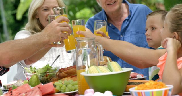 Multi-generational family enjoying outdoor brunch together, raising glasses of orange juice in a toast. Perfect for themes of family bonding, healthy eating, outdoor activities, summer gatherings, or intergenerational relationships. Useful for advertisements, articles, and websites promoting family values, health, and lifestyle.