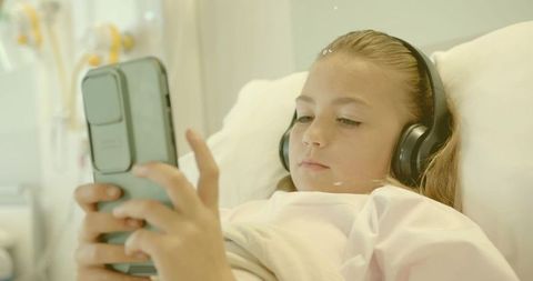 Child resting in hospital bed listening to smartphone audio with headphones during recovery