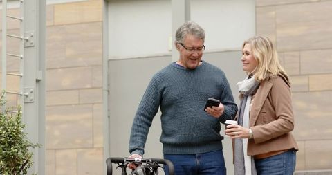 Mature couple sharing phone and coffee while strolling with bicycle through urban plaza