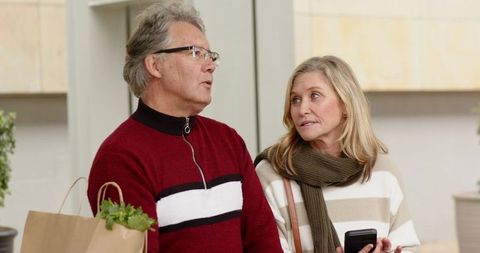 Senior couple strolling through shopping district holding groceries and checking smartphone