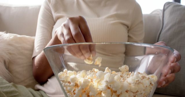 Close-up of a person grabbing popcorn from a bowl while sitting on a couch. The image evokes a sense of relaxation and comfort, ideal for use in content related to home leisure, casual food consumption, and cozy home settings. Perfect for advertisements promoting snacks, home entertainment, or cozy living.