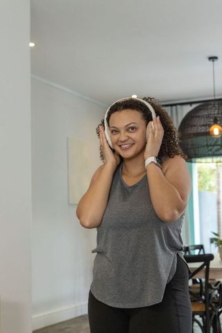 African american woman smiling while listening on white headphones and smartwatch at home