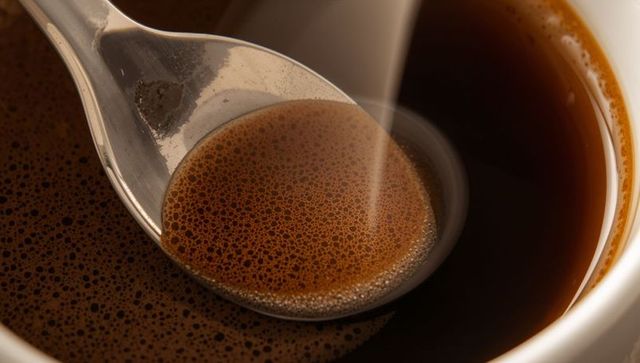 Macro metal spoon holding espresso crema with rising steam and glossy coffee reflections
