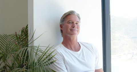 Serene senior man leaning on white wall by window with indoor plant in minimalist home