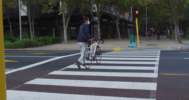 Man walking bicycle across zebra crossing in urban setting while wearing a protective mask. Ideal for showcasing city transportation, pedestrian safety, COVID-19 precautions, and active city lifestyle.