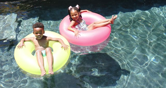 Children are floating in colorful water tubes in a pool, enjoying a sunny summer day. Ideal for ads related to summer activities, family vacations, and pool safety. This can also be used for promoting outdoor recreational products and children's swimwear.