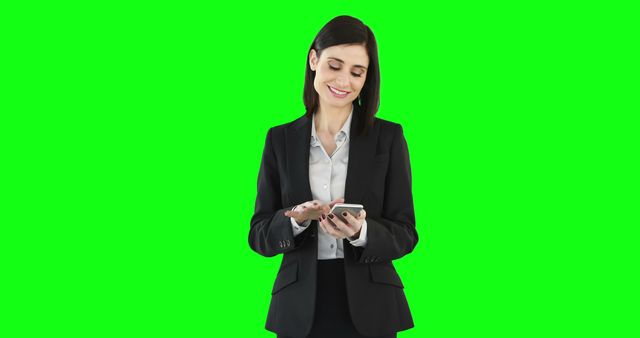 Businesswoman in black suit smiling while using smartphone. The green screen background can be easily replaced for diverse settings. Ideal for marketing campaigns, promotional materials, business presentations, and advertisements highlighting professionalism and modern technology in the workplace.