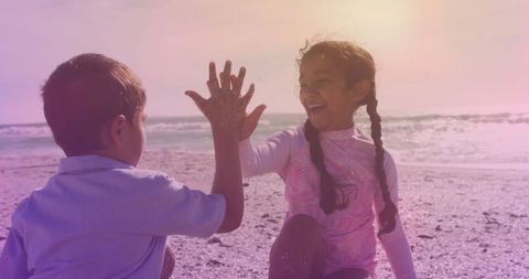 Two Hispanic children stand on a beach, smiling and sharing a joyous high five. The scene captures a sense of carefree fun amid sun and sand, symbolizing a strong sibling bond. The light creates a soft ambiance. Ideal for concepts of family togetherness, joyful childhood memories, or promoting events like National Siblings Day or family vacations.