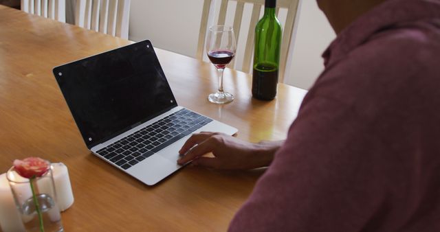 Man sitting at wooden table working on laptop with wine glass and candle. Suitable for promoting remote work, home office setups, or relaxation environments.