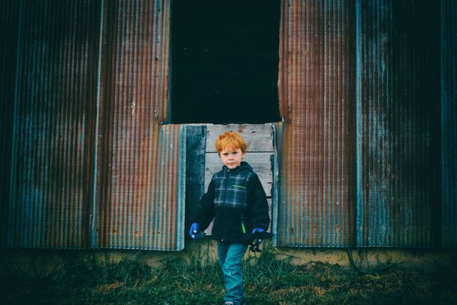 A child with red hair is standing in the entrance of a barn. He is wearing a jacket and blue gloves. The barn has a corrugated metal exterior. This image can be used to depict rural life, childhood, outdoor activities, or part of an agricultural theme.