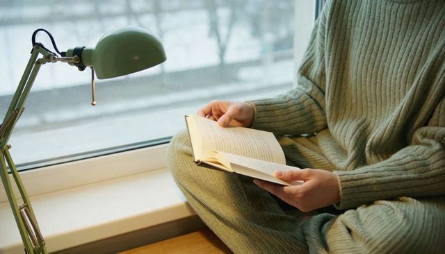 Reading in ribbed sage knit on wide window sill under green pullchain lamp cozy winter nook