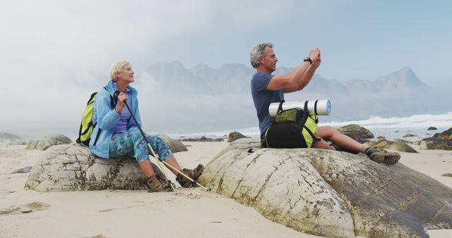 Senior couple hiking on a beach with mountainous background. Man taking photos with camera, woman resting with walking sticks and backpack. Ideal for themes of active aging, adventure trips, outdoor activities, and travel promotions.