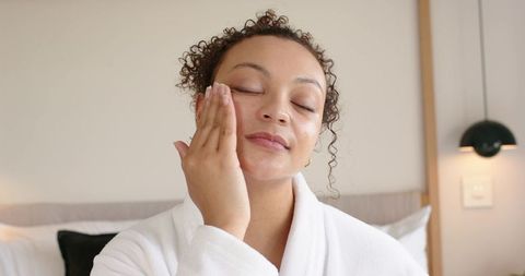 African american woman applying facial moisturizer in robe cozy bedroom morning routine