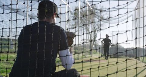 Sunlit catcher receiving pitch in netted cage while teammate practicing pitching on turf