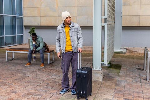 Diverse male travelers waiting with suitcase in city plaza wearing beanie and headphones