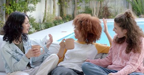 Diverse women chatting poolside on outdoor sofa holding ceramic mugs casual lifestyle