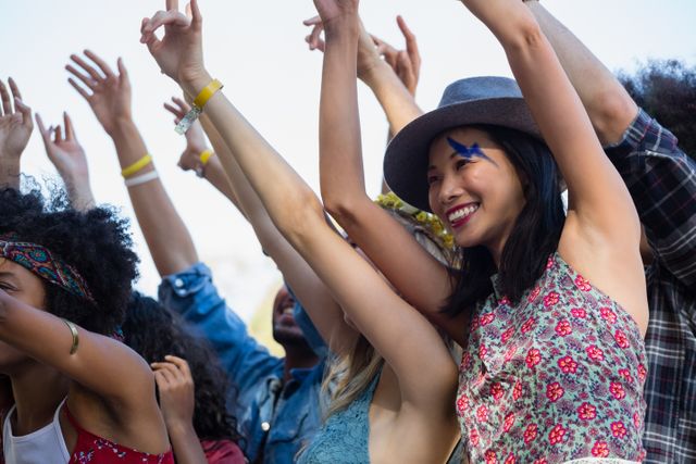Group of diverse female friends raising their hands and smiling at an outdoor music festival. Ideal for use in advertisements, social media posts, and promotional materials related to music events, festivals, outdoor activities, and youth culture.