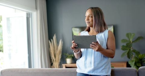 Mature woman holding tablet and smartwatch in bright modern living room, neutral decor