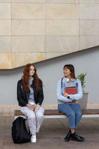 Two female students sitting on bench sharing coffee and notebooks on campus