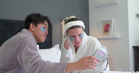 Couple enjoying at-home spa with under-eye patches, bathrobes and handheld mirror
