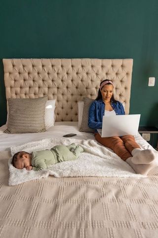 Working mother on bed with sleeping newborn, laptop remote work and cozy morning routine