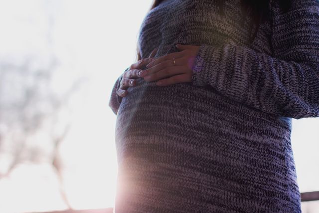 Pregnant woman with long hair in a gray knit sweater gently holding her pregnant belly. Soft natural light and backlighting create a serene and calm atmosphere. Suitable for maternity care products, parenting blogs, family-focused advertising, prenatal care features, and motherhood-related content.