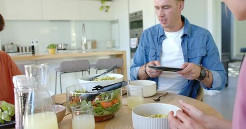 Family dining with man using tablet at modern kitchen table featuring salad and pasta bowls