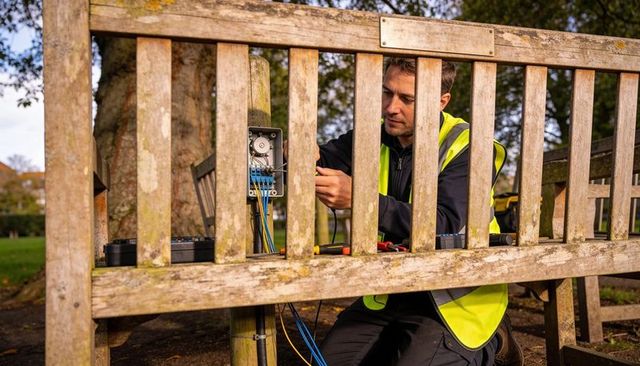Kneeling technician repairing park bench electrical junction box with blue cables