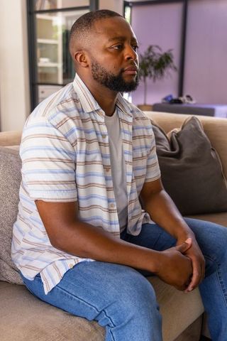 Contemplative african man sitting on beige sofa holding clasped hands in living room