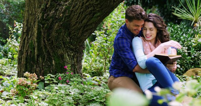 Couple enjoying time together, lounging under a tree in a lush garden setting. Both appearing relaxed and happy while man hugs woman who is holding a book. Ideal for use in romantic contexts, relaxation messages, relationship and lifestyle promotions, or depicting harmony and peaceful moments in nature.