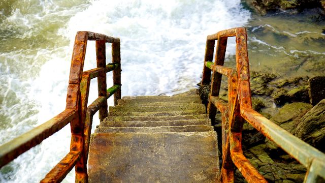 A rusty staircase descends towards the crashing ocean waves, highlighting the effects of weather and marine conditions. Suitable for themes of coastal erosion, nature's power, and nautical environments. Ideal for travel blogs, environmental awareness campaigns, or maritime-themed designs.