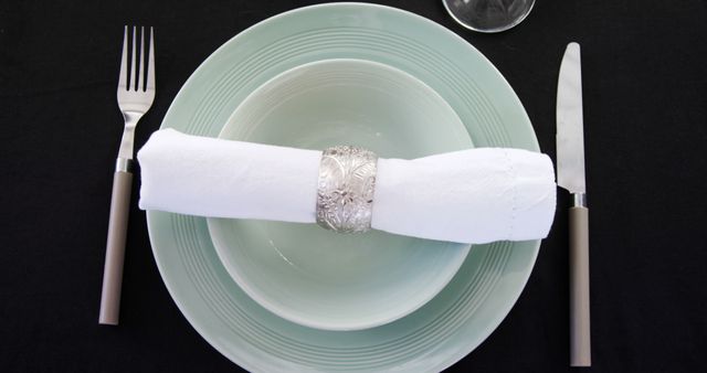 An elegant table setting features a neatly rolled white napkin secured with a decorative napkin ring, centered on a pastel blue plate, flanked by a fork on the left and a knife on the right. The simplicity and symmetry of the arrangement convey a sense of formality and readiness for a dining occasion.