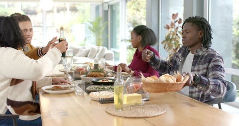 Diverse friends sharing meal and passing basket of rolls around bright modern dining table