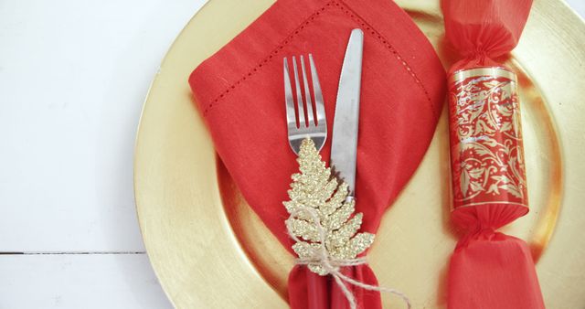 A festive table setting features a gold charger plate paired with a red napkin, elegant cutlery, and a decorative gold leaf, with copy space. The arrangement suggests a holiday or special occasion meal, emphasizing a sense of celebration and elegance.
