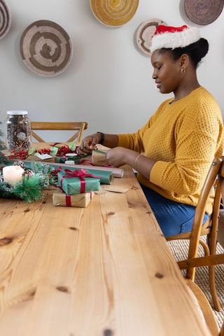 Holiday gift wrapping by african american woman in santa hat on cozy wooden dining table
