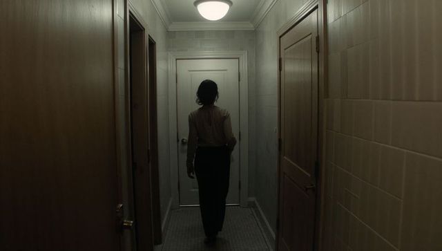 Woman walking down dim corridor toward closed white door moody cinematic suspense shadowy
