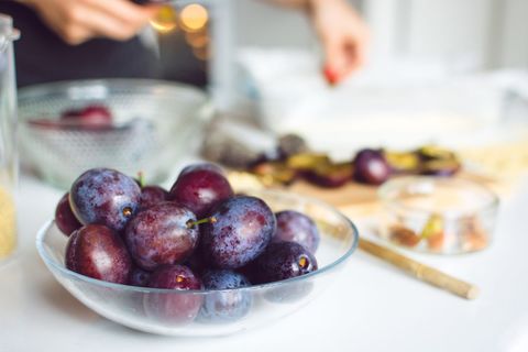 Displaying ripe purple plums in glass bowl with hands prepping fruit on wooden board