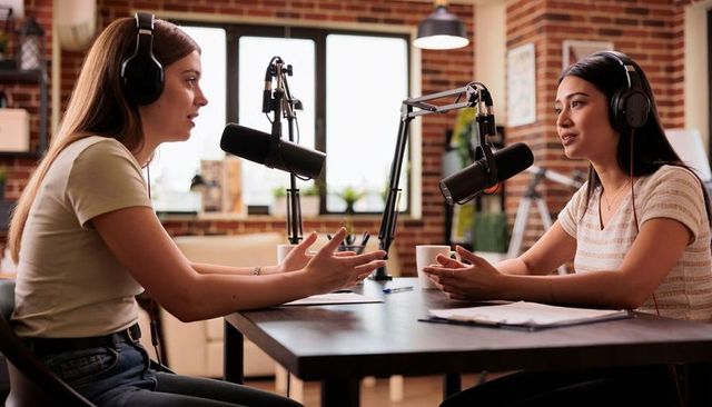 Two women recording conversational podcast in cozy brick studio with broadcast microphones