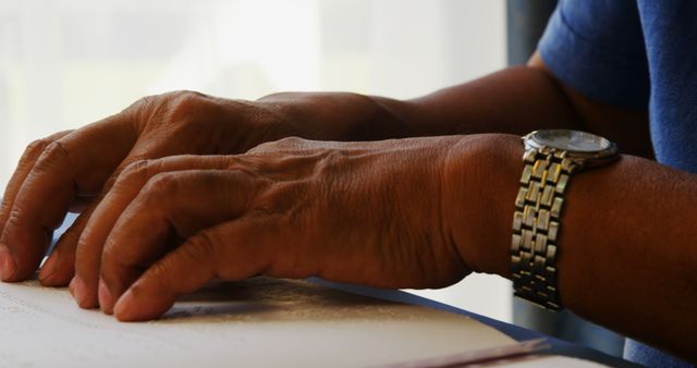 A close-up view highlights the hands of a senior individual, reading a book or document, with copy space. The presence of a wristwatch suggests an attention to time or schedule during this quiet moment of reading or studying.