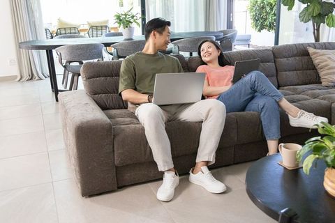 Diverse couple working remotely on laptop and tablet relaxing on cozy gray sectional sofa