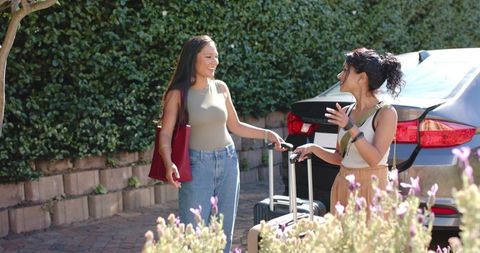 Sunlit diverse female friends talking on driveway with suitcases by silver sedan road trip