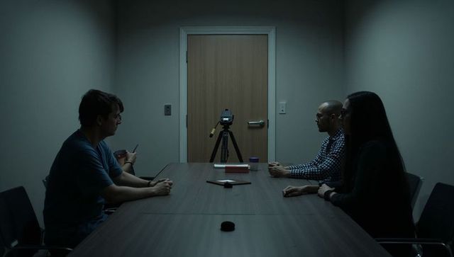 Three colleagues sitting in dim meeting room facing tripod camera for interview recording