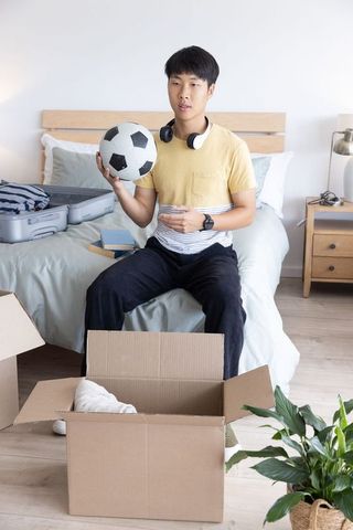 Asian man unpacking for move holding soccer ball on bed with suitcase boxes and headphones